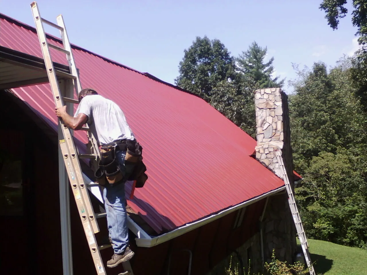 Roofer on a ladder beside a red metal roof and stone chimney — Soffit & Fascia Repair by Roofing Brothers in Hopatcong